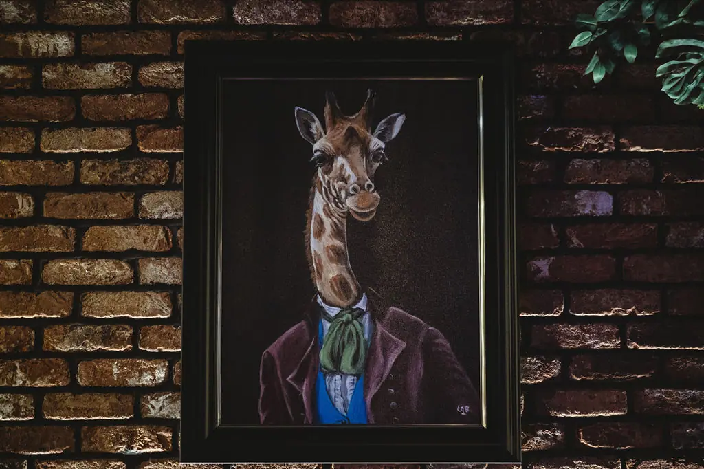 Framed painting of a giraffe in a velvet jacket on a brick wall